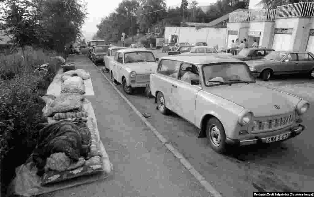 Уцекачы з Усходняй Нямечыны сьпяць побач са сваімі аўтамабілямі Trabant у Будапешце. З драматычным паваротам Вугоршчыны да свабоды ў 1989 годзе тысячы ўсходніх немцаў, якія пакутавалі ад пастаяннага сачэньня з боку тайнай паліцыі Штазі, пачалі рух на ўсход. Вугоршчына была адной з «брацкіх сацыялістычных краін», у якія ўсходнім немцам дазвалялася падарожнічаць.